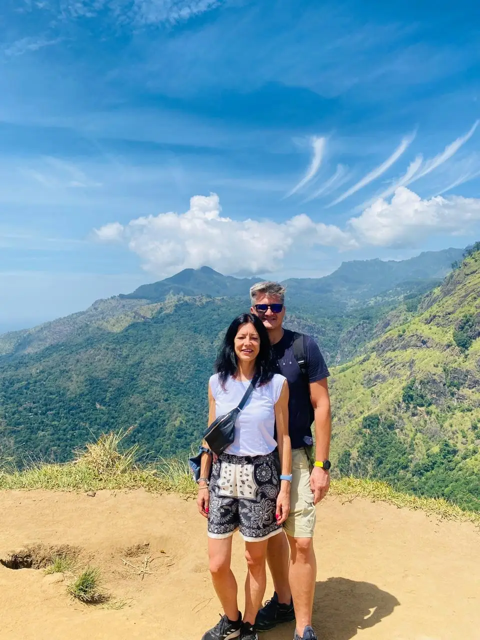 A couple at a mountain lookout under a wide blue sky streaked with high cloud