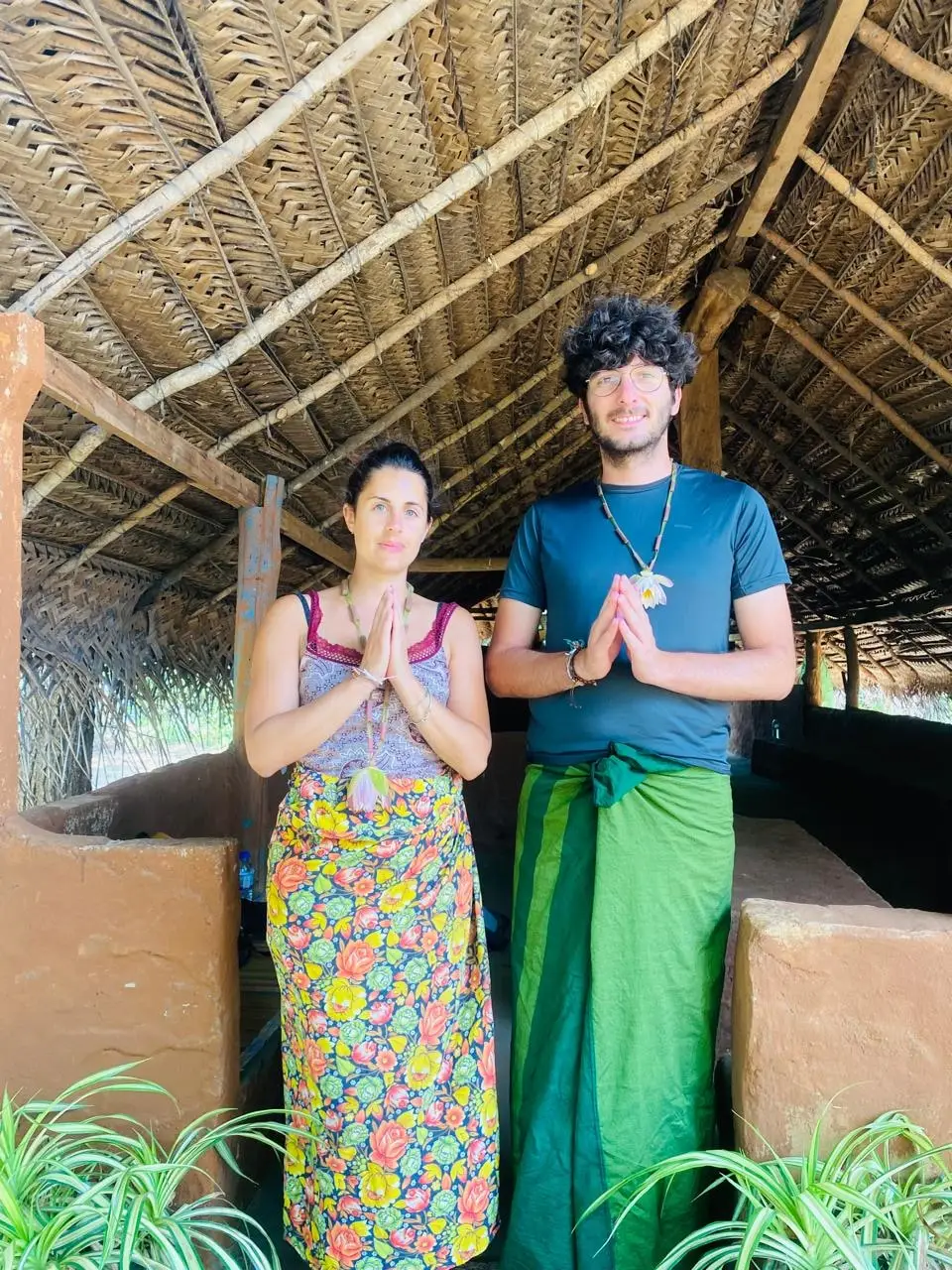 Two guests wearing traditional sarongs, hands pressed together in greeting, under a thatched roof